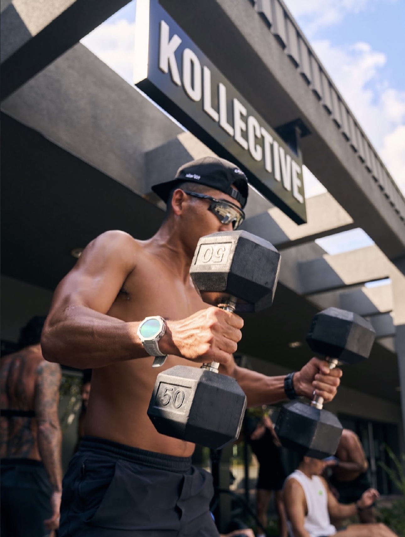Member training outside the Kollective with 50lb dumbbells, club signage overhead